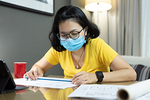 Asian female Architect wear eyeglasses using scale ruler for measurement on her blueprints during quarantine in pandemic Coronavirus. Young Woman interior designer in yellow shirt, blue surgical mask working from home during Covid 19.